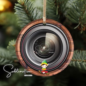 以下が含まれることがあります： カメラレンズの形をした木製のクリスマスオーナメントで、中に漫画の妖精が入っています。オーナメントは茶色で、黒いレンズと赤い縁取りがあります。妖精は緑色の帽子と赤と白の服を着ています。