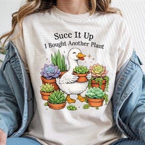 May include: Cream-colored t-shirt with a graphic of a smiling white duck holding a potted succulent. The text "Succ It Up I Bought Another Plant" is above the duck. Several potted succulents in various colors surround the duck.