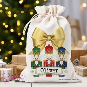 Puede incluir: Una bolsa de regalo crema con cordón con tres soldados cascanueces con uniformes verdes, rojos y azules. Un gran lazo dorado está encima de los soldados. El nombre "Oliver" está impreso en una pancarta.