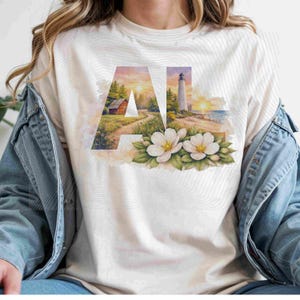 May include: Cream-colored t-shirt featuring the letters "AL" with a watercolor design. The "A" depicts a cabin and path, while the "L" shows a lighthouse. White flowers and green leaves are at the bottom.