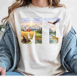 May include: Cream-colored t-shirt featuring the letters "MT" with nature scenes. The "M" displays a bear and cub, a red barn, and mountains. The "T" shows an eagle and a fish jumping out of the water.