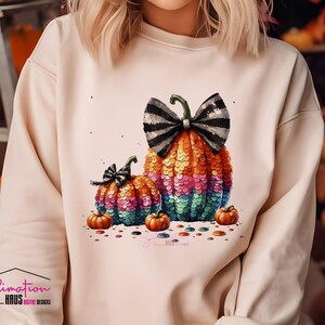 Puede incluir: Una sudadera blanca con un diseño de calabaza de lentejuelas de colores. La calabaza tiene un lazo blanco y negro. Hay calabazas más pequeñas a cada lado de la calabaza grande.