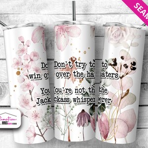 May include: White tumbler with a floral design and the text "Don't try to win over the haters. You're not the jackass whisperer." Features pink flowers, gold accents, and a stainless steel straw. The word "Seamless" is in the top right corner.