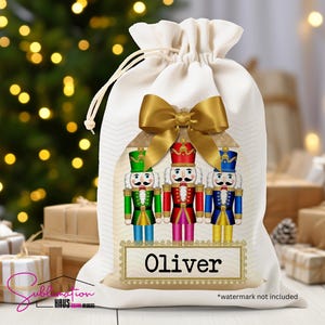 May include: A white drawstring gift bag with a gold bow and three nutcracker figures. The bag has a wooden-style nameplate that reads "Oliver". The nutcrackers are in green, red, and blue uniforms.
