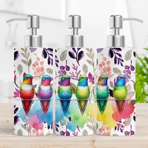 May include: Three colorful hummingbird soap dispensers with a floral and watercolor design. The dispensers are white with a silver pump.