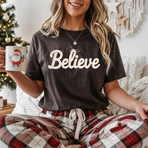 May include: Dark gray t-shirt with the word "Believe" in a cream-colored, textured font. The shirt is paired with red and white plaid pajama pants. A white mug with a Santa Claus graphic is held in the left hand.