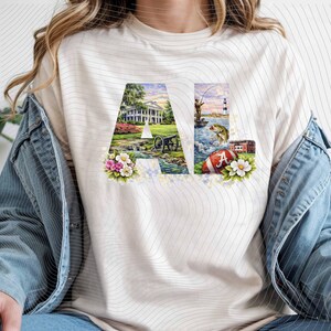 May include: Cream-colored t-shirt featuring the letters "AL" with colorful illustrations. The "A" displays a white house, cannon, and flowers. The "L" shows a fisherman, lighthouse, football, and a building.