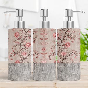May include: Three soap dispensers with silver pumps. They have a floral design with pink roses and brown stems on a beige background. The lower section has a grey, textured, wood-like appearance.