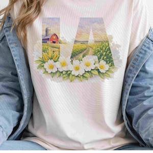 Puede incluir: Camiseta blanca con las letras "IA" en un diseño de acuarela. La "I" muestra un granero rojo y un silo, mientras que la "A" representa un camino rural a través de un campo de maíz. Flores blancas y hojas verdes adornan la parte inferior de las letras.