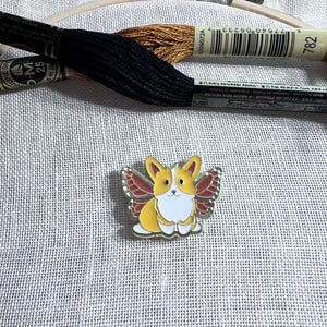 May include: A gold-colored enamel pin featuring a cartoon corgi with red butterfly wings. The corgi is yellow with white markings and has a white belly. The pin is on a white fabric background.