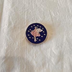 May include: A round, blue enamel pin with a gold border. The pin features a white fairy with pink wings and a pink dress, flying against a background of gold stars.