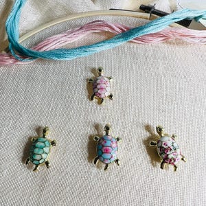May include: Four gold-colored turtle charms with different floral patterns on a white fabric background. The turtles are in a row, with the top turtle having a pink and white floral pattern, the second turtle having a blue and white floral pattern, the third turtle having a green and white floral pattern, and the fourth turtle having a pink and white floral pattern.
