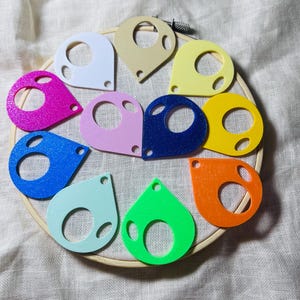 May include: A collection of eleven teardrop-shaped plastic yarn bobbins in various solid colors including white, beige, yellow, orange, green, light blue, blue, dark blue, pink, magenta, and lavender, arranged in a circle on a wooden embroidery hoop.