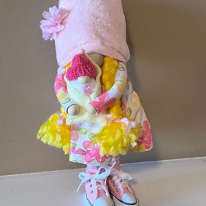 May include: A pink and yellow gnome doll wearing a pink hat and holding a smaller pink and white gnome doll. The larger gnome is wearing a floral dress and pink sneakers.