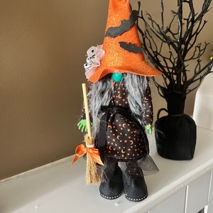 May include: A Halloween witch gnome figurine. The gnome wears a tall orange glitter hat with black bat cutouts, a black dress with orange crescent moons and stars, and black boots. It holds a broom with an orange bow.