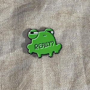 May include: A green frog-shaped enamel pin with the word "REALLY?" in white. The pin has a glossy finish and black details on the eyes. The pin is set against a textured, neutral-colored fabric.