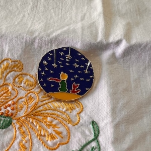 May include: A gold-colored enamel pin featuring a blue background with a depiction of The Little Prince and his fox looking up at a starry night sky.