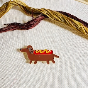May include: A gold-colored dachshund pin with a red hot dog and yellow mustard on its back.