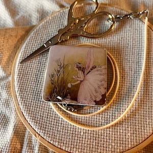 May include: A gold pair of embroidery scissors with a decorative handle and a small charm with a fairy illustration. The charm is attached to a white cord.