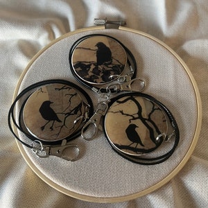 May include: Three round black and white keychains with a silhouette of a bird on each. The keychains are attached to black cords and silver clasps. The keychains are displayed on a wooden embroidery hoop.