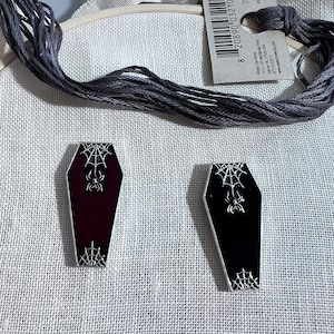 May include: Two enamel pins shaped like coffins with a spider and web design. One coffin is black and the other is red.