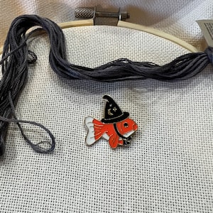 May include: A red and white fish wearing a black witch hat with a crescent moon and star design. The fish is on a white Aida cloth with a gray embroidery floss.