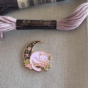 May include: A gold-tone enamel pin featuring a white cat sleeping on a crescent moon with pink and green floral accents.
