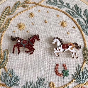 May include: Two horse-shaped enamel pins, one burgundy and one white with brown and black details, are displayed on a cream-colored fabric. The pins are surrounded by embroidered cacti, stars, and a horseshoe.