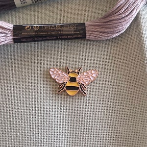 May include: A gold-coloured bee pin with black stripes and pink wings. The bee is on a light grey fabric background.