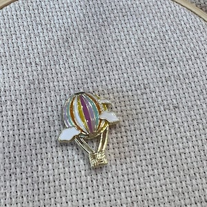 May include: A gold-colored hot air balloon pin with a striped pattern in pink, yellow, and blue. The balloon has white clouds attached to it.