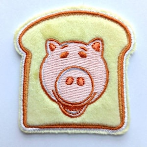 Könnte beinhalten: Ein gestickter Aufnäher aus gelbem Filz mit einem Schweinegesicht in einer Scheibe Brot. Das Schwein ist rosa mit orangefarbenen Details und das Brot ist orange umrandet.