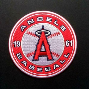Puede incluir: Un parche bordado en rojo, blanco y azul con las palabras "Angels Baseball 1961" alrededor de una pelota de béisbol blanca con una "A" roja con un halo arriba.
