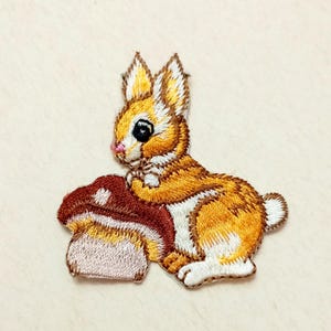 Puede incluir: Parche bordado con un conejo de dibujos animados en tonos naranja, blanco y marrón, sosteniendo una seta. El conejo tiene una nariz rosa y un ojo negro. El parche está diseñado para su aplicación en ropa o accesorios.