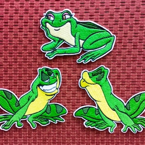 May include: Three green frog embroidered patches with white outlines. The frogs are in different poses, one is sitting with its arms crossed, one is sitting with its legs crossed, and one is standing with its arms outstretched.