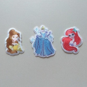Puede incluir: Tres parches bordados para planchar con princesas Disney: Bella con cabello castaño y un vestido amarillo, Cenicienta con cabello azul y un vestido azul, y Ariel con cabello rojo y un top de concha púrpura.