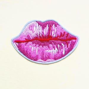 May include: Embroidered patch featuring a pair of glossy, pink lips with a red lip line. The lips are outlined in a light blue border. The patch is designed to be ironed on or sewn onto clothing or accessories.
