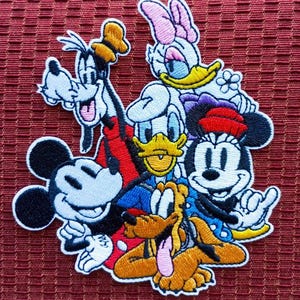 Peut inclure: Écusson brodé représentant Mickey Mouse, Minnie Mouse, Donald Duck, Daisy Duck, Dingo et Pluto. L'écusson a une bordure blanche et est posé sur un fond rouge. Les personnages sont dans leurs styles de dessins animés classiques, avec des couleurs vives.
