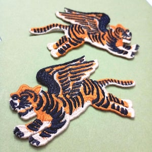 May include: Two embroidered patches featuring tigers with wings. The tigers have orange and black stripes, with black and orange wings. The patches are on a light green background.