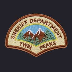 Puede incluir: Un parche bordado marrón y beige con el texto "Sheriff Department Twin Peaks" en letras amarillas. El parche presenta una imagen estilizada de dos montañas con picos nevados y árboles verdes.