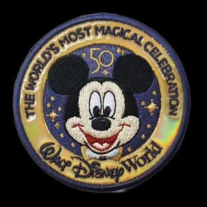 Könnte beinhalten: Ein gestickter Aufnäher in Gold und Blau mit Mickey Mouse, der einen Zylinder trägt. Der Aufnäher trägt den Text "The World's Most Magical Celebration" und "Walt Disney World" am Rand. Die Zahl 50 befindet sich in der Mitte des Aufnähers.