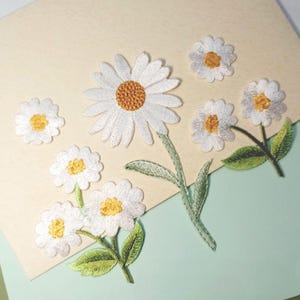 May include: Embroidered daisy patches in various sizes. The white petals surround a yellow center, with green stems and leaves. The patches are arranged on a light beige and light green background.