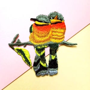 May include: Embroidered patch depicting two vibrant birds on a branch with a leaf. The birds display yellow, orange, and green feathers, with grey heads and black beaks. The branch and leaf are green and yellow. The patch is set against a pink and yellow backdrop.