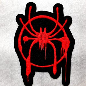 Puede incluir: Un parche bordado negro que presenta un diseño de araña roja estilizada encerrado dentro de un borde circular. El bordado rojo incluye efectos de pintura que gotea en las patas de la araña y el círculo, creando una apariencia texturizada.
