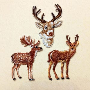 Op de afbeelding: Drie geborduurde hertenpatches in bruin- en wittinten. Eén patch is een hertenkop en de andere twee zijn herten met een volledig lichaam. De patches hebben gedetailleerde stiksels en staan op een lichtbeige achtergrond.