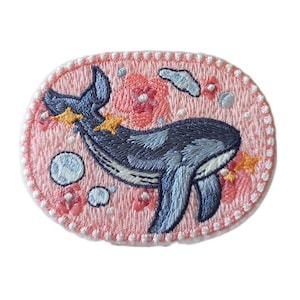 May include: A pink embroidered patch featuring a blue whale swimming in a field of pink flowers and white stars. The whale is surrounded by white bubbles.