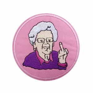 May include: A pink and white embroidered patch featuring a cartoon of an older person with grey hair wearing glasses and a purple sweater, giving the middle finger.
