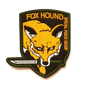Könnte beinhalten: Gestickter Aufnäher mit einem gelben Fuchskopf mit Messer, auf einem schwarzen und grünen Schild. Der Text "FOX HOUND SPECIAL FORCE GROUP" ist oberhalb und rechts vom Fuchs angeordnet.