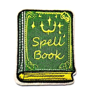 Puede incluir: Un parche bordado con forma de libro rectangular con cubierta verde oscuro, detalles cosidos en amarillo y el texto Spell Book en cursiva amarilla. El diseño presenta estrellas amarillas y un borde inferior amarillo.