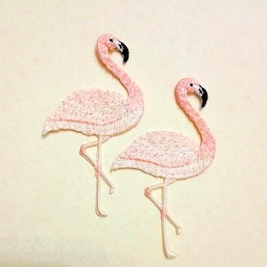 Pode incluir: Dois emblemas bordados de flamingos com detalhes em fio rosa claro, pernas longas e finas e bicos pretos. As aves estão representadas de perfil, exibindo costuras intrincadas que definem as asas, os pescoços e os corpos dos flamingos.