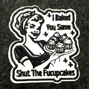 Könnte beinhalten: Schwarz-weißer Aufnäher mit einer Frau im Retro-Stil, die Cupcakes serviert. Der Text lautet "I Baked You Some" und "Shut The Fucupcakes". Der Aufnäher hat einen weißen Rand und ist zum Aufnähen geeignet.